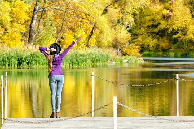 Ragazza in cappello in piedi sul molo con una mano sollevata autunno soleggiato vista posteriore