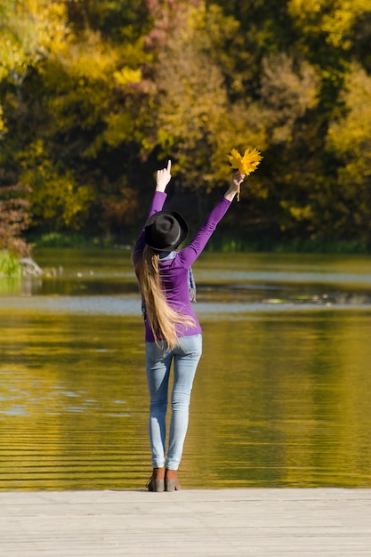 Girl in hat standing on the pier with leaves in hands and points up. Autumn sunny day. Back view