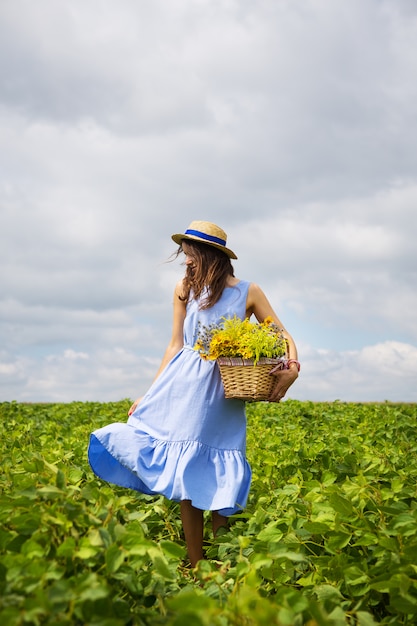 Girl in a hat stands on a green field with a basket of flowers