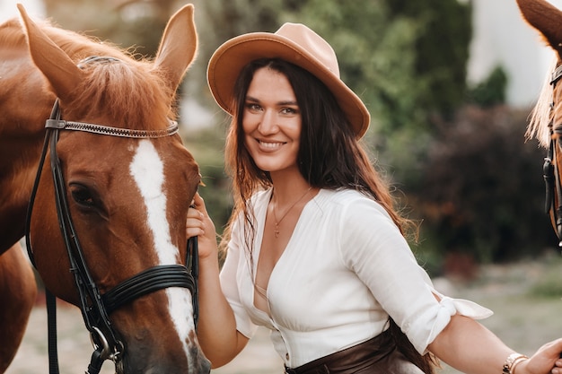 A girl in a hat stands next to horses in nature.