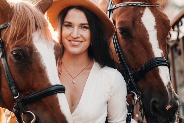 A girl in a hat stands next to horses in nature