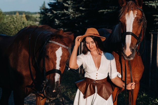 A girl in a hat stands next to horses in nature