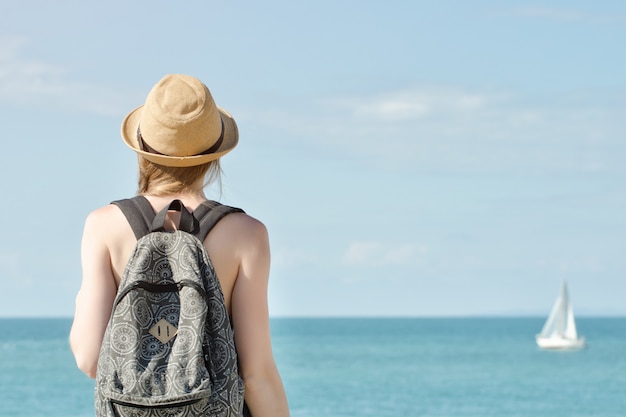 Девушка в шляпе с рюкзаком, стоя на береговой линии. Парусник на расстоянии. Вид со спины