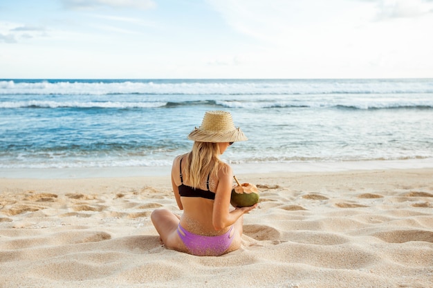 Girl in a hat with a coconut sits on the beach, back view