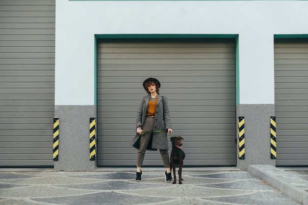 Girl in a hat and with curly hair holds a leash with a dog in her hand and poses at the camera