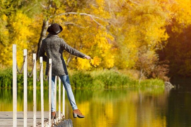 Girl in a hat with leaves in hands standing on the dock. Autumn, sunny. Back view