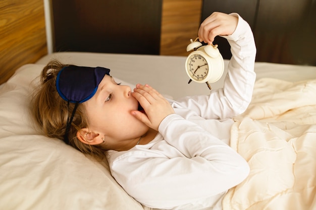 Photo girl hates getting up early in the morning the blonde holds the alarm clock yawning in bed covers her mouth with her hand