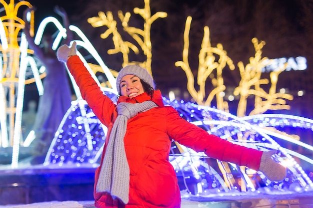 Photo girl having fun on christmas decoration lights street. young happy smiling woman wearing stylish knitted scarf and jacket outdoors. model laughing. winter wonderland city scene, new year party.