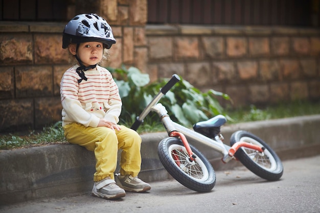 Girl in a helmet is sitting on the curb with a running bike lying next to her.