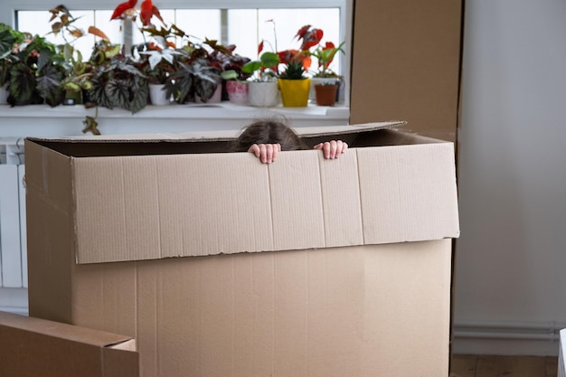 The girl hides in a big box and looks out of it funny in a room inside the home Moving to a new house unpacking things renting an apartment mortgage housing issue