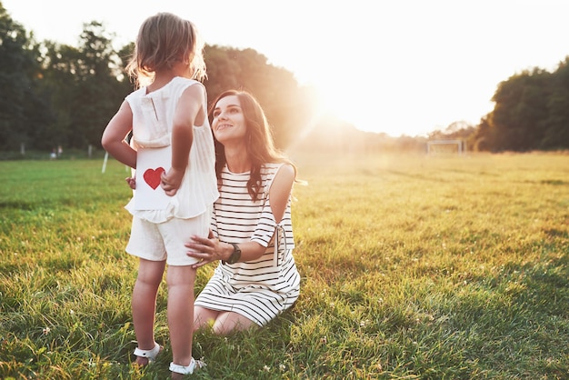 Girl hides the book with red heart on the back from the smiling sitting mother