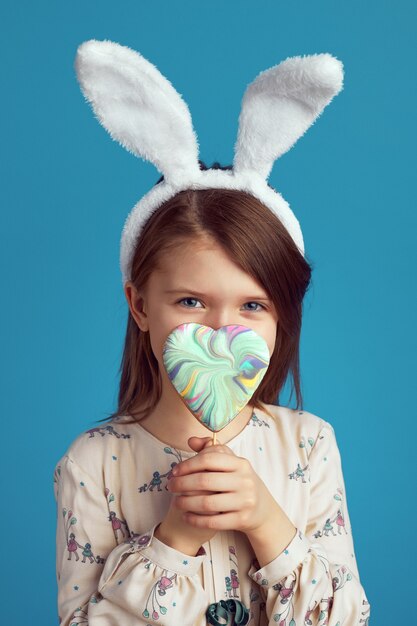 Girl hiding behind a heart shaped cookie and wearing bunny ears over blue