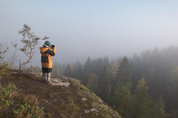 Girl hiker on a rocky hill above a foggy forest takes a photo