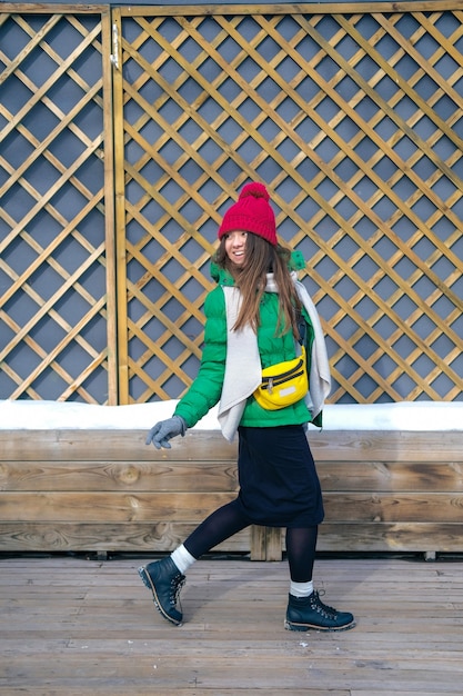 Girl hipster in bright clothes laughing and smiling on the wall of wooden boards. Young happy woman in a red knit hat, green jacket, grey scarf, yellow bag. Women's fashion. Autumn,spring trend