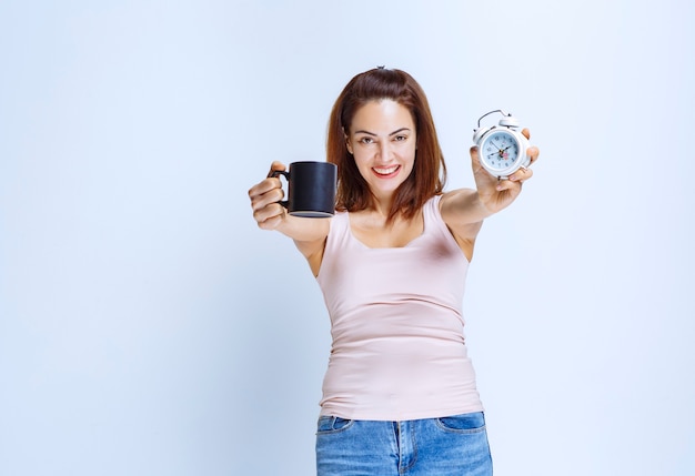 Girl holding an alarm clock in one hand and a black coffee mug in another. 