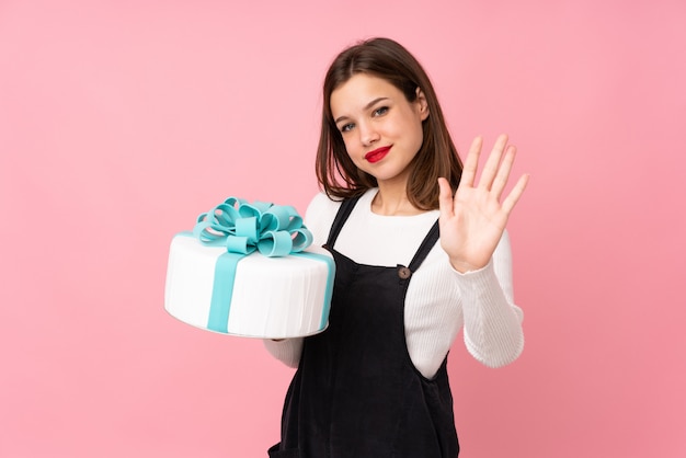 Girl holding a big cake on pink saluting with hand with happy expression