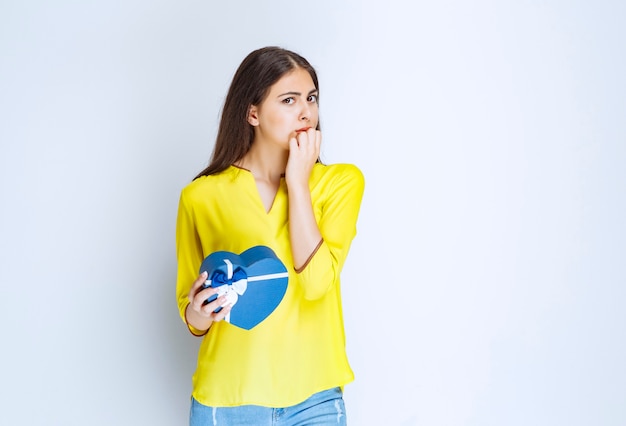 Girl holding a blue heart shape gift box and thinking or hesitating.