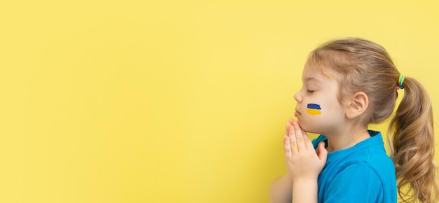 Girl holding folded hands in prayer with yellow and blue colors of the Ukrainian flag on her cheek Copy space