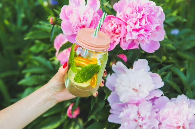 Girl holding fresh lemonade in jar with straw. Hipster summer drink in hand with peonies. Eco-friendly in the nature. Lemons, oranges and berries with mint in the glass. Beautiful spring flowers.