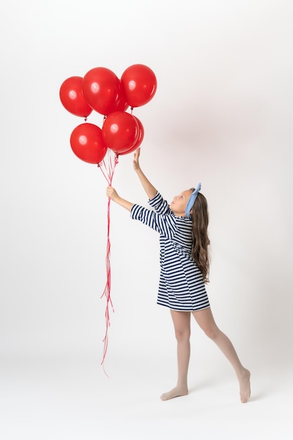 Девушка держит в руке красные воздушные шары, другая рука тянется к воздушным шарам и стоит на цыпочках на белом