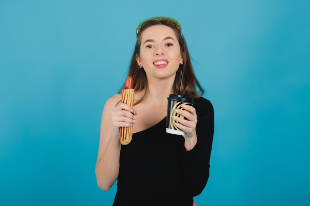 girl holding hot dog and glass with coffee on blue background