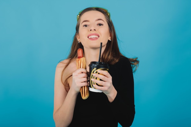 girl holding hot dog and glass with coffee on blue background