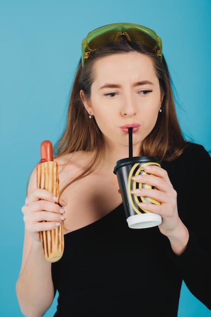 girl holding hot dog and glass with coffee on blue background