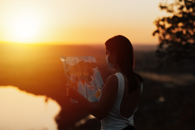 Girl holding map world, she wearing a mask on sunset surface. Tourist girl exploring route.