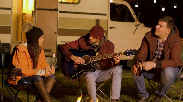 마시멜로를 들고 있는 소녀와 그의 친구 중 한 명이 가을의 추운 밤에 캠프 파이어 주변에서 기타를 치며 노래하고 있습니다. 산에서 캠핑.