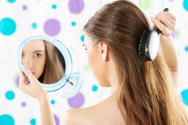 Girl holding a mirror in which looks and combing her long brown hair. The girl is photographed from the rear.