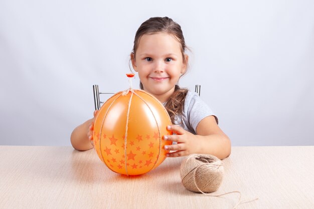 Girl holding an orange balloon wrapped with jute rope, DIY pumpkins for Halloween, self-isolation hobby.