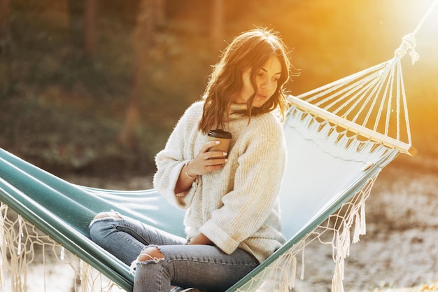 Girl holding a paper cup of coffee in the park on a hammock