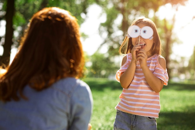 Girl holding paper mask on the face and showing to her mother in the park