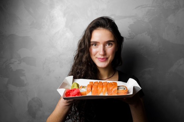 Girl holding philadelphia rolls in a paper box on gray background. Sushi, food delivery.