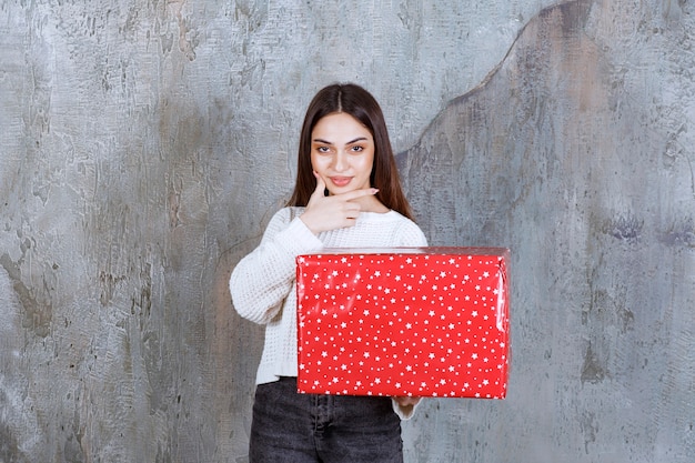 Girl holding a red gift box with white dots on it and looks thoughtful and dreamy.