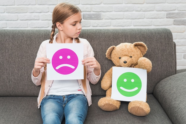 Photo girl holding sad smileys paper looking at teddy bear with happy smileys