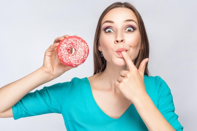 Ragazza che tiene e mostra una ciambella rosa che si succhia il dito con grandi occhi su sfondo grigio