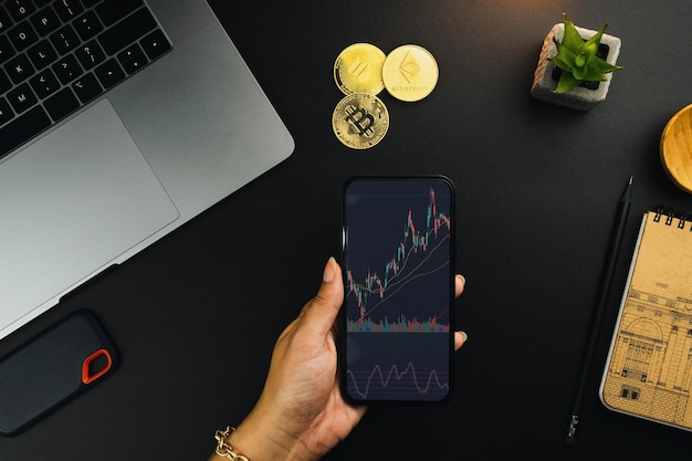 Girl holding a smartphone with financial stock market graph on the screen on black table with a cryptocurrencies coins and computer Office environment Stock Exchange