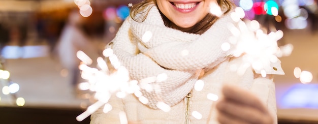 Girl holding a sparkler in her hand. Outdoor winter city background, snow, snowflakes.