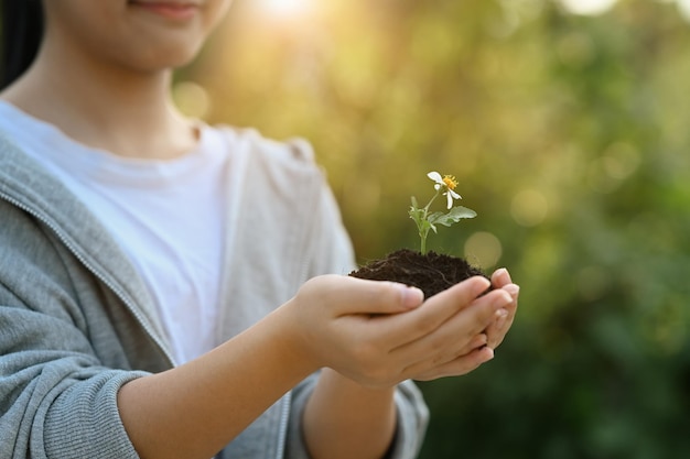 ぼやけた緑の自然の背景と日光に対して若い植物を手に持っている女の子アースデイエコロジーコンセプト