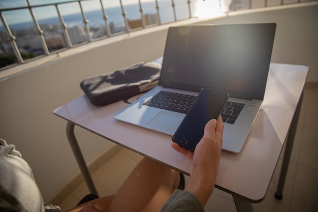 Girl holds her smartphone in hand while working with her laptop sitting in the balcony Digital nomad