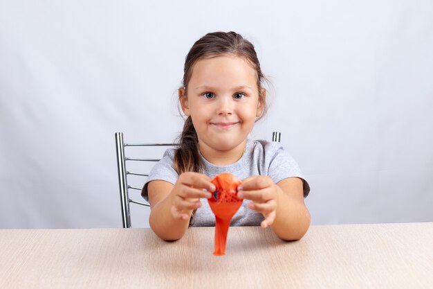 The girl holds an inflated balloon, smiles and sits at a table on a white background.