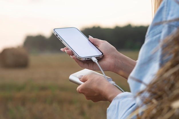 Girl holds a mockup of a smartphone with a white screen in her hands. Power Bank charges the phone against the background of nature.