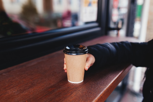 Girl holds a paper cup of coffee in hand near cafe. 