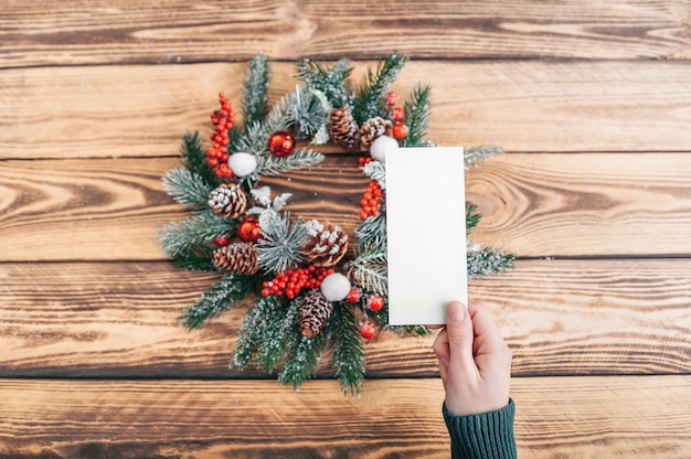 The girl holds a postcard on the background of a christmas wreath and a wooden table. layout. place to insert.