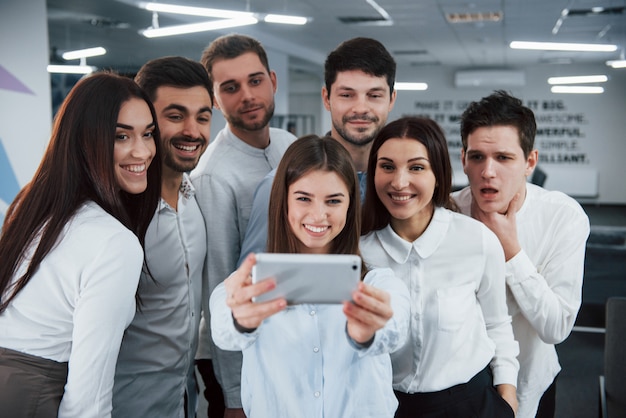 La ragazza tiene il telefono color argento. giovane gruppo che fa selfie in vestiti classici nell'ufficio illuminato buono moderno