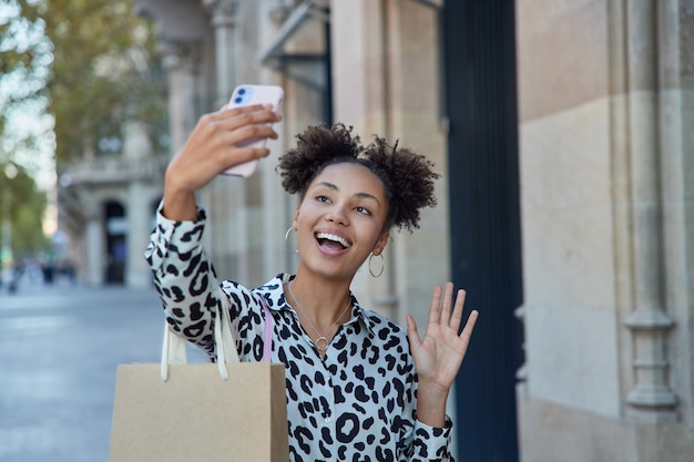 girl holds smartphone waves hello makes online call communicates with friend carries paper bag returns from mall has good time