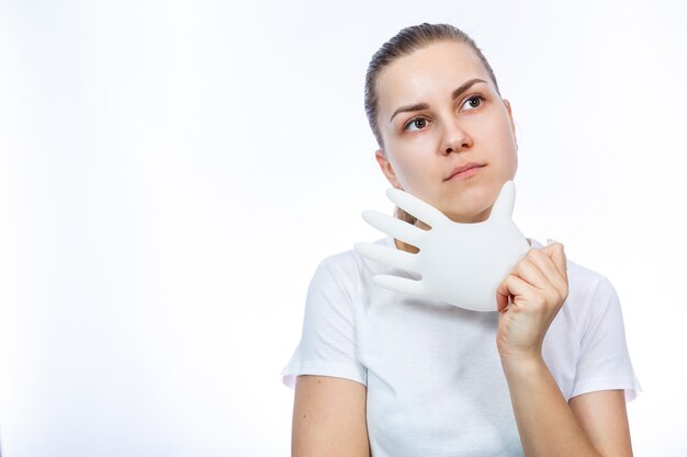 The girl holds white medical gloves in her hands. Protection against germs and the virus. She is in a white T-shirt on a white background.