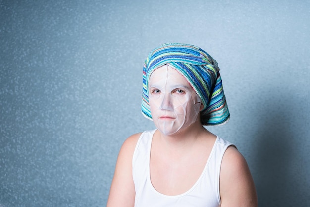 A girl in home clothes and with a towel on her head does a cosmetic procedure at home using a sheet mask for facial skin care