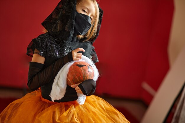 Girl at home in halloween costume with pumkin Jack or Laurent in hands, Child wearing black face mask protecting from coronavirus, halloween in quarantine